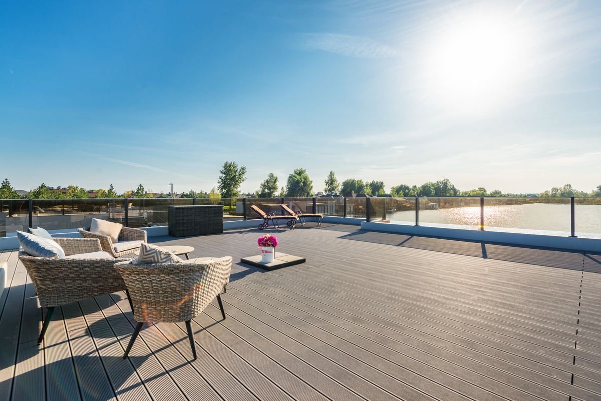 Spacious modern composite deck with gray grooved boards, wicker lounge furniture, and a glass railing overlooking a lake on a sunny day