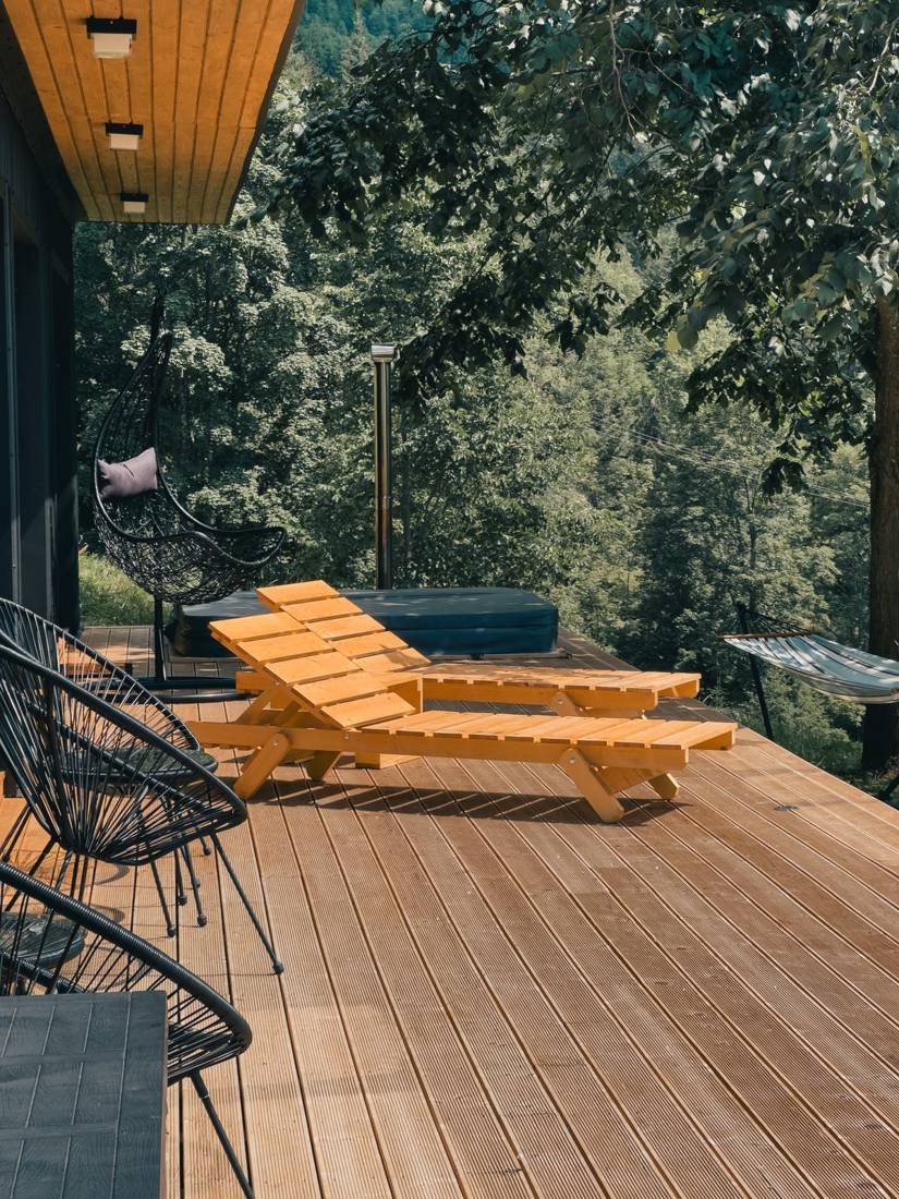 Warm-toned wooden deck with two slatted lounge chairs and a hanging egg chair, surrounded by lush green trees