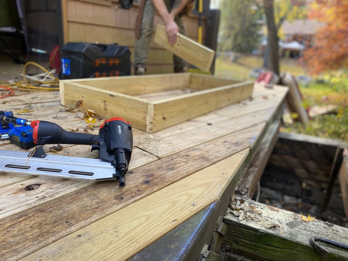 Deck under construction with a framing nailer resting on fresh pressure-treated boards while a carpenter carries lumber in the background