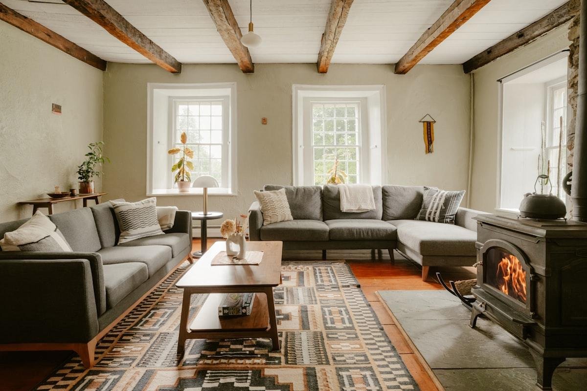 Cozy farmhouse living room with dark exposed ceiling beams, gray sofas, a patterned rug, and a lit wood-burning stove
