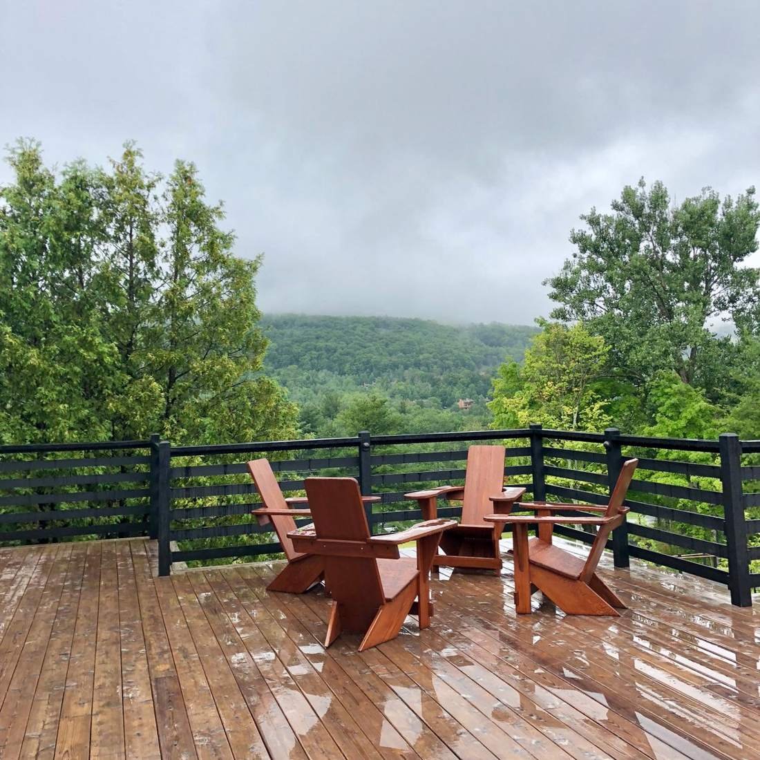 Wooden deck with Adirondack chairs and a dark horizontal-rail fence overlooking forested mountains under low clouds
